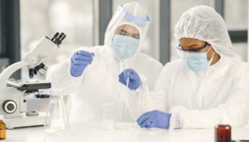 Pesquisadores em laboratório realizando experimentos ou análises clínicas. Eles utilizam equipamentos de proteção individual (EPIs) completos, manuseando amostras líquidas com pipetas e equipamentos de análise, como um microscópio de bancada.