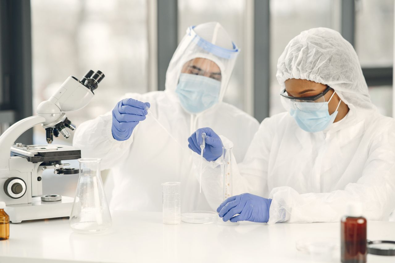 Pesquisadores em laboratório realizando experimentos ou análises clínicas. Eles utilizam equipamentos de proteção individual (EPIs) completos, manuseando amostras líquidas com pipetas e equipamentos de análise, como um microscópio de bancada.